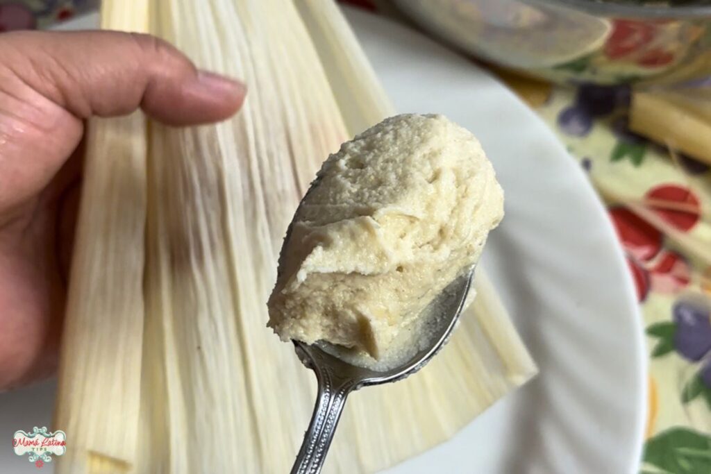 A person is holding a spoon full of homemade tamale masa dough with one hand and a corn husk with the other.