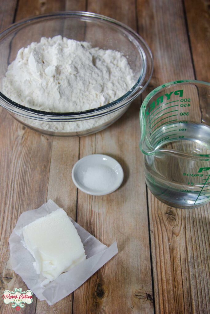 Ingredientes de tortillas de harina: harina, sal, manteca y taza medidora con agua sobre mesa de madera.