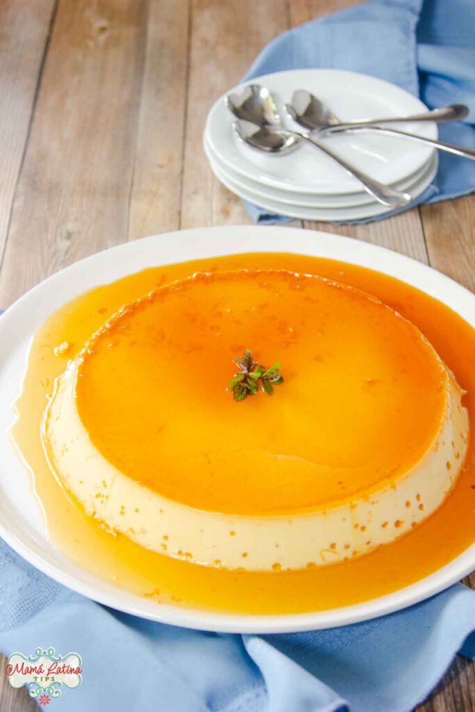 Un flan de vainilla con caramelo encima de un plato blanco, junto a una torre de tres platos. 