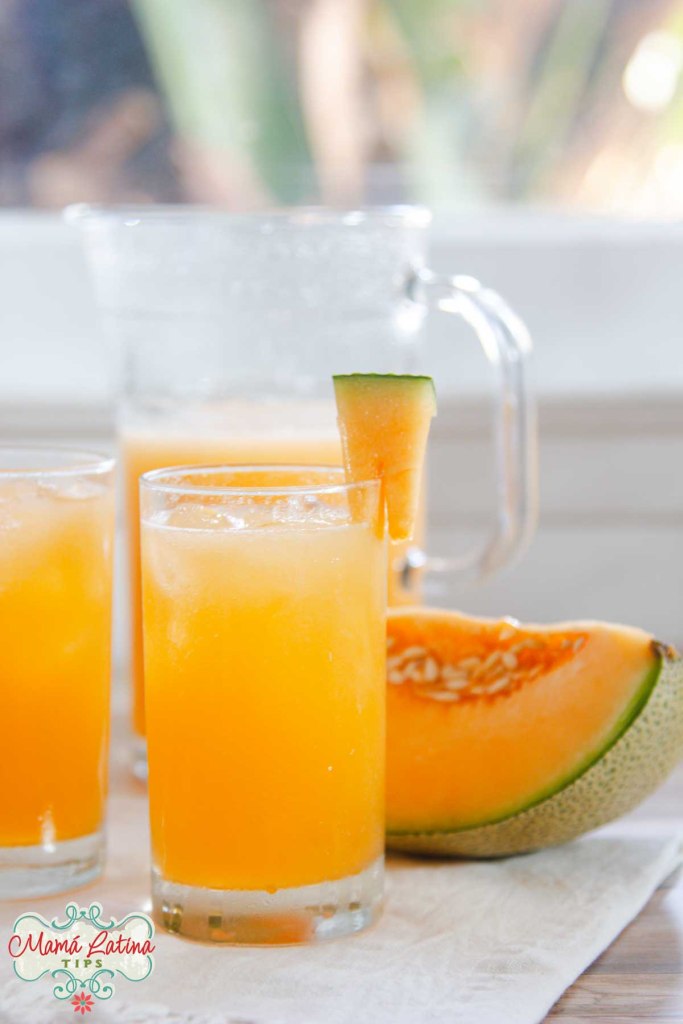 Two glasses of agua fresca de melon on a table with a slice of cantaloupe on one glass and a pitcher in the background.