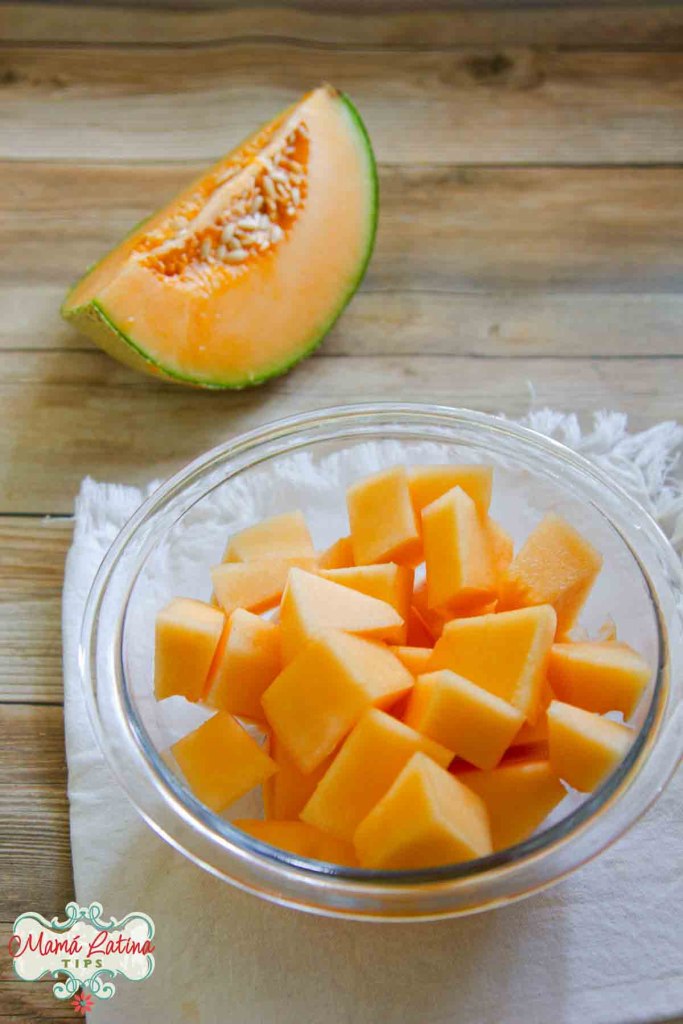 A glass bowl filled with diced cantaloupe on a wooden table, with a sliced cantaloupe wedge in the background.