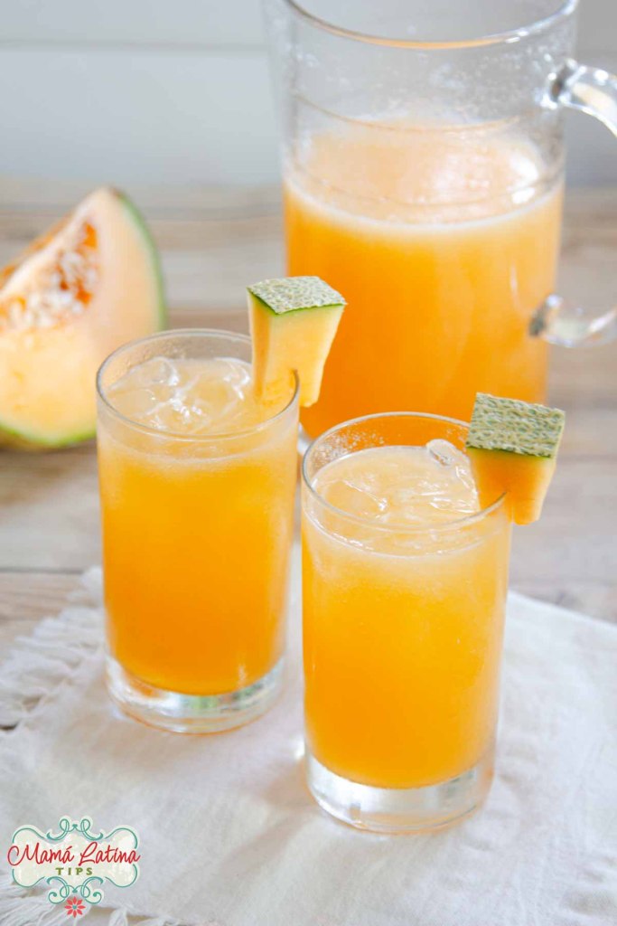 A pitcher and two glasses filled with cantaloupe agua fresca on a wooden table, garnished with cantaloupe slices.