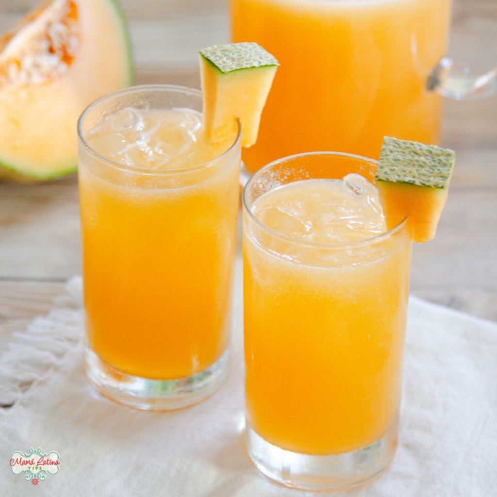 Two glasses of cantaloupe agua fresca with ice, garnished with melon slices on a wooden table.