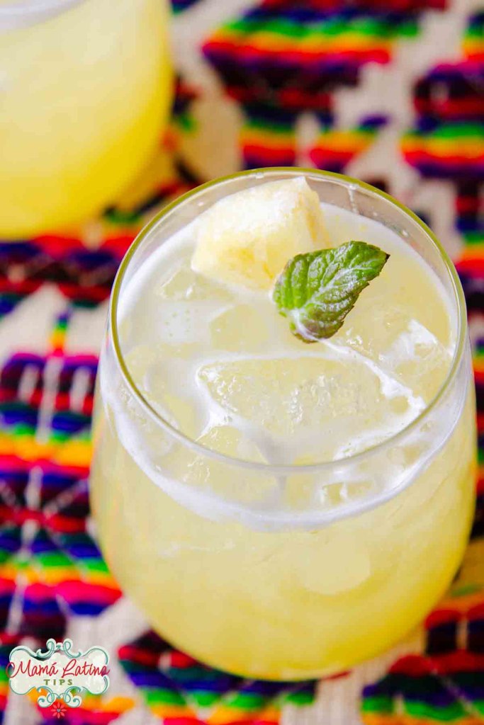 A glass of pineapple agua fresca with ice and a mint leaf on a colorful woven cloth.