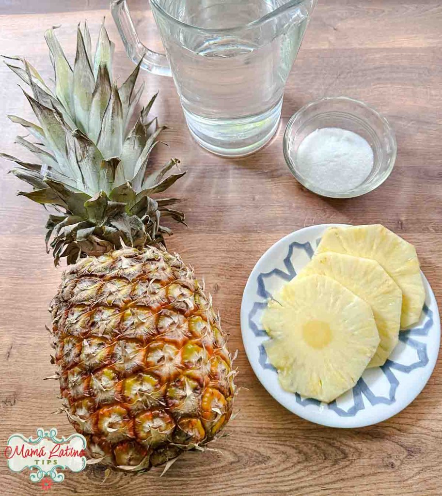 A fresh pineapple beside sliced pineapple on a plate, with a glass of water, jug, and a bowl of sugar on a wooden table.