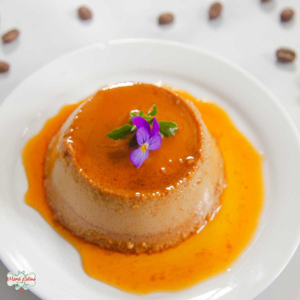 Coffee flan dessert on a white plate, garnished with a purple flower and surrounded by coffee beans.