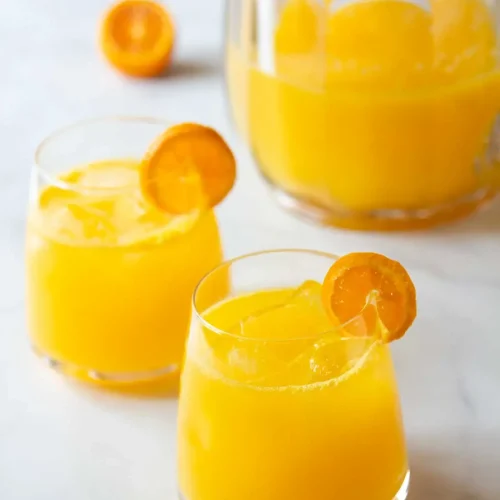 Two glasses of mandarin agua fresca with ice and orange slices, next to a pitcher filled with more agua fresca.
