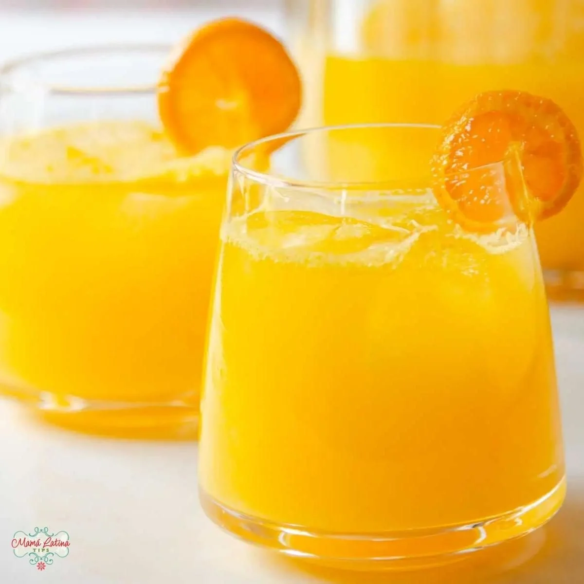 Two glasses of refreshing mandarin orange agua fresca with ice, each garnished with an orange slice, are shown on a white surface—perfect for a festive touch.