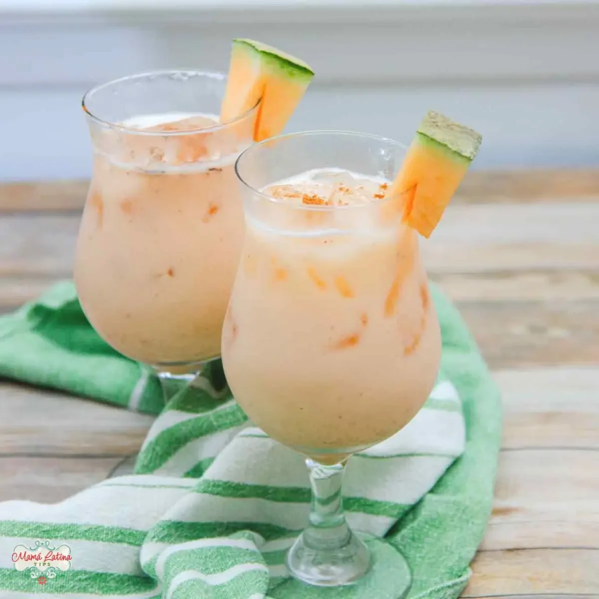 Two iced glasses of creamy cantaloupe drink, each garnished with a cantaloupe wedge, sit on a green and white striped towel on a wooden surface.