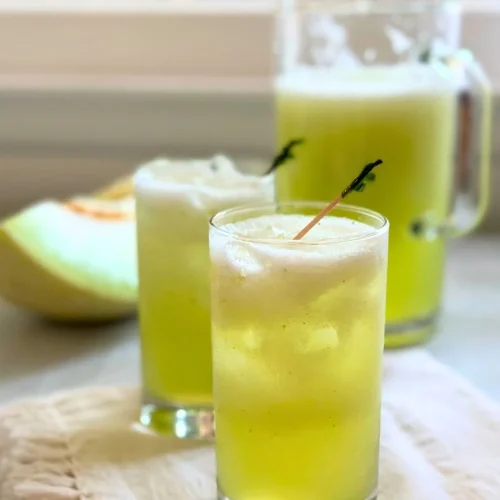 Two glasses and a pitcher filled with refreshing Honeydew agua fresca, topped with foam, sit on a light-colored cloth with a melon slice in the background—an inviting honeydew drink perfect for your next agua fresca recipe.