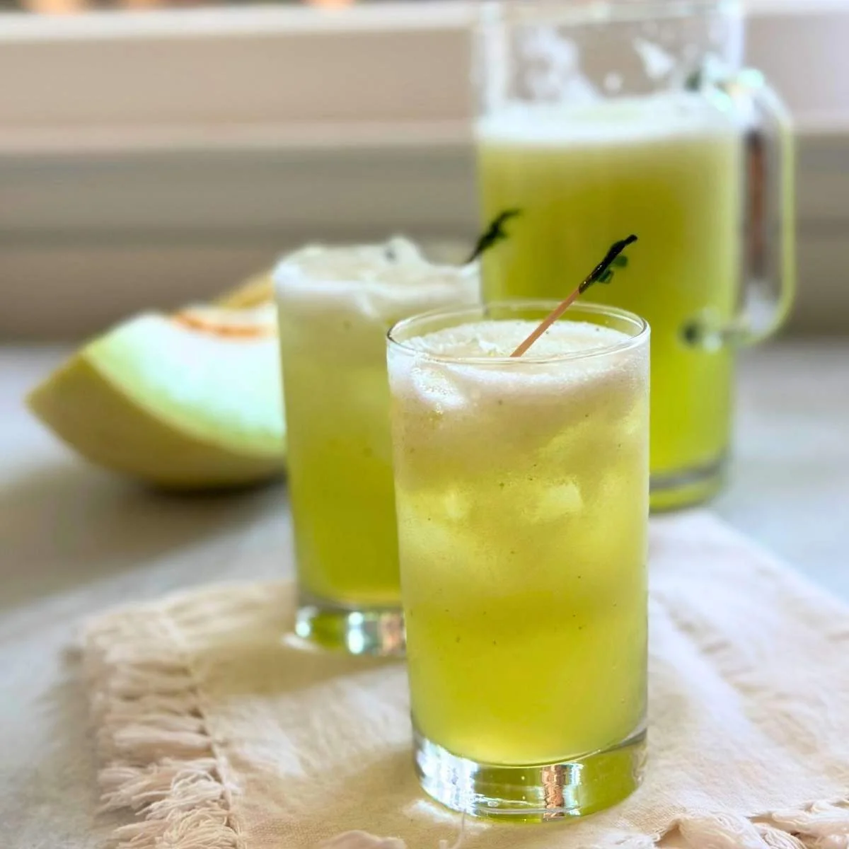 Two glasses and a pitcher filled with frothy, light green honeydew agua fresca sit on a cloth, with a wedge of melon in the background.