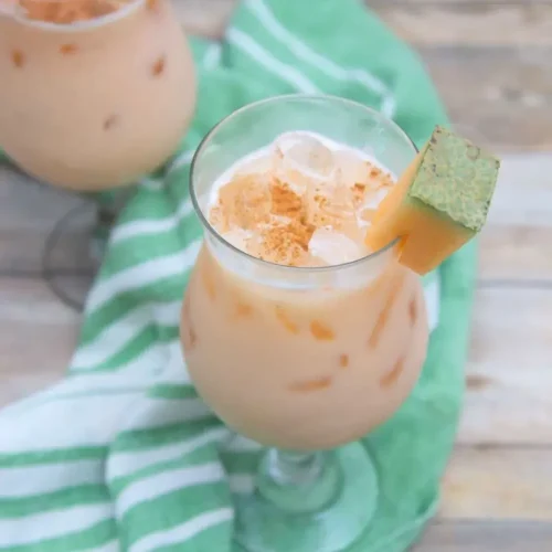 Dos vasos de agua fresca de melón con hielo, adornados con gajos de melón y espolvoreados con canela, sobre una superficie de madera con un paño de rayas verdes debajo.