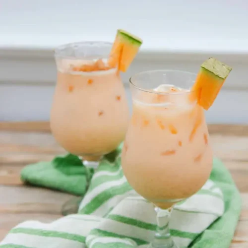 Two glasses of iced cantaloupe agua fresca, made with a refreshing recipe, each garnished with a cantaloupe slice, sit on a green and white striped towel on a wooden surface.