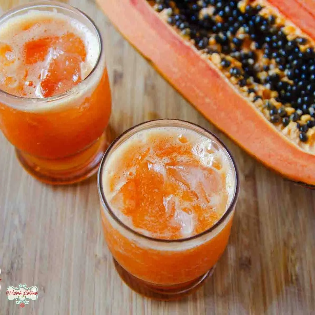Two glasses of refreshing papaya drink with ice sit on a wooden surface next to a halved papaya filled with seeds, perfect for enjoying as an agua fresca.