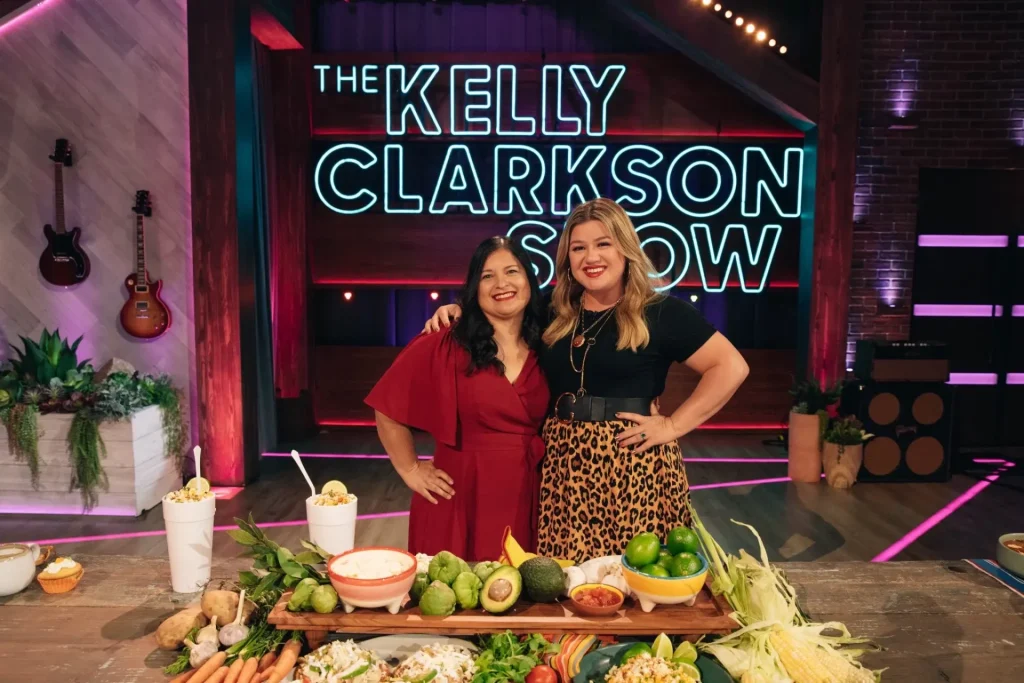 Silvia from Mama Latina Tips and another woman stand smiling in front of a table filled with various fruits and vegetables on the set of The Kelly Clarkson Show.