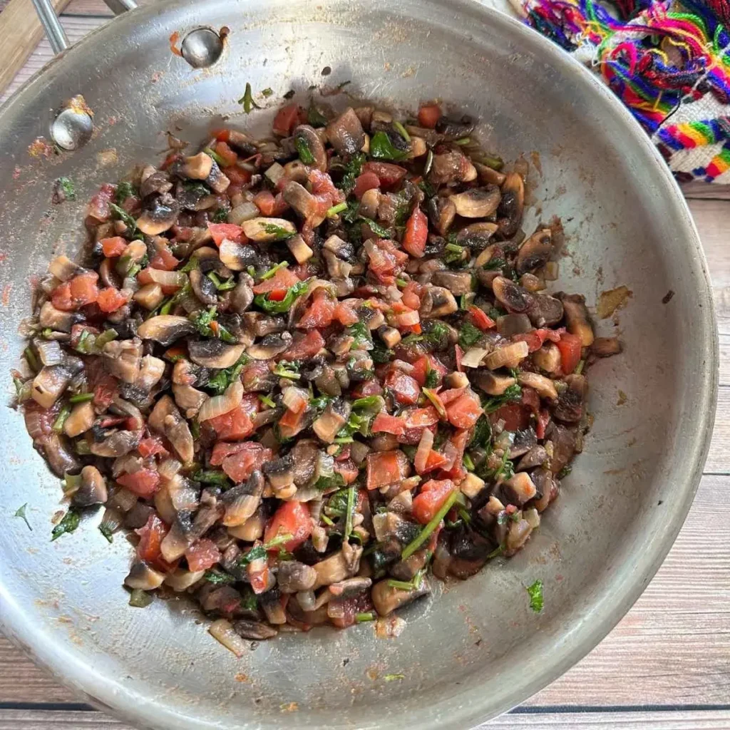 A stainless steel pan containing a mixture of sautéed mushrooms, diced tomatoes, and chopped herbs on a wooden surface.