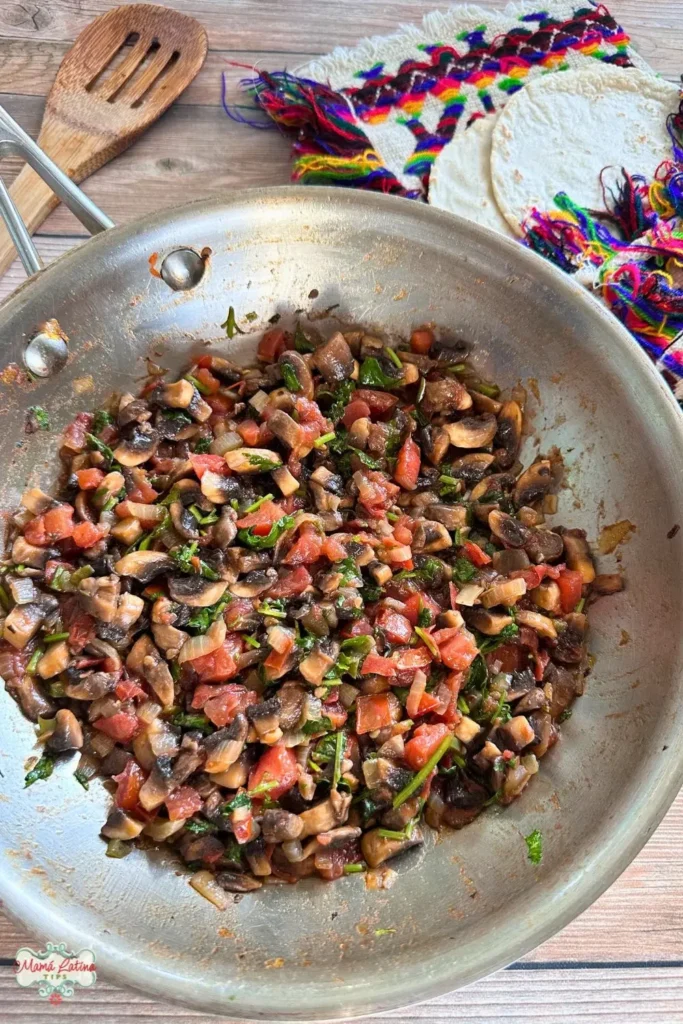 A skillet with sautéed mushrooms, tomatoes, and herbs sits on a wooden surface next to corn tortillas and a colorful woven cloth.