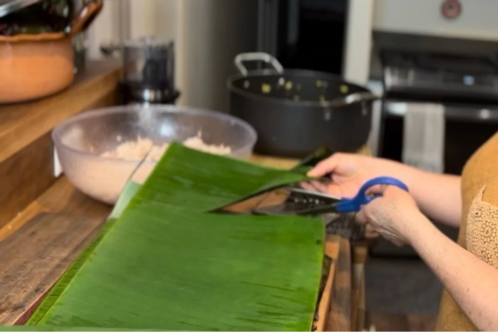 Una persona cortando hojas de plátano con tijeras sobre una mesa de madera en la cocina. En la parte trasera hay ollas, cazuelas y un ball con masa para tamales