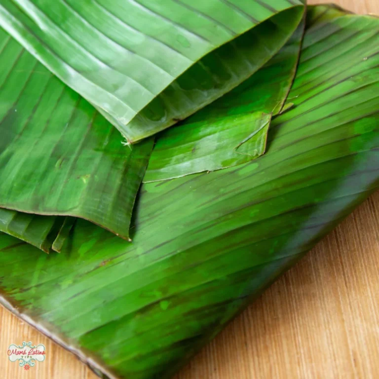 Several green banana leaves are stacked on a wooden surface, perfect for learning how to prepare banana leaves for cooking or tamales