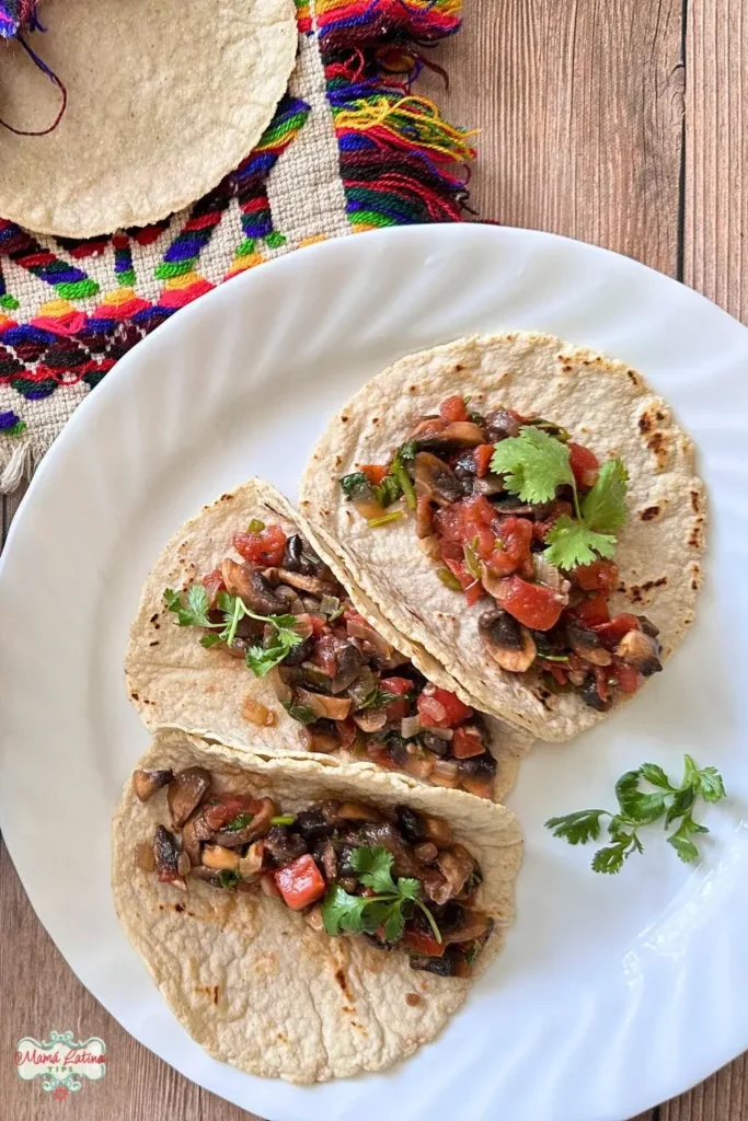 Three soft tacos filled with Mexican style mushrooms, tomatoes, and herbs are arranged on a white plate, garnished with fresh cilantro, on a wooden surface.