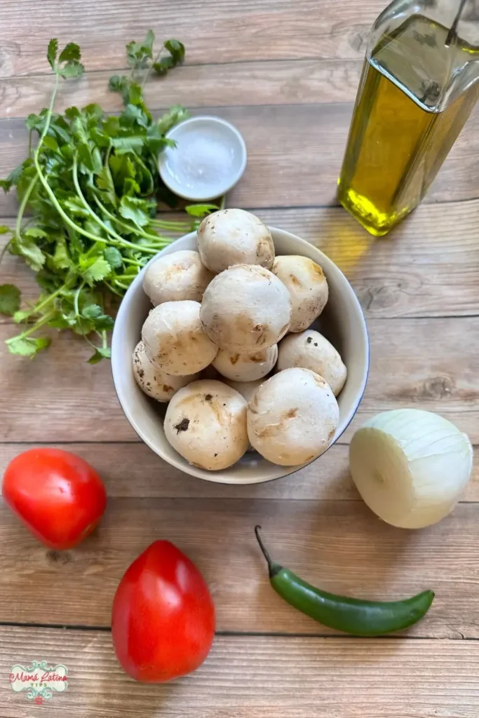 A bowl of white mushrooms surrounded by cilantro, a small dish of salt, a bottle of olive oil, a whole onion, two tomatoes, and a green chili pepper on a wooden surface.