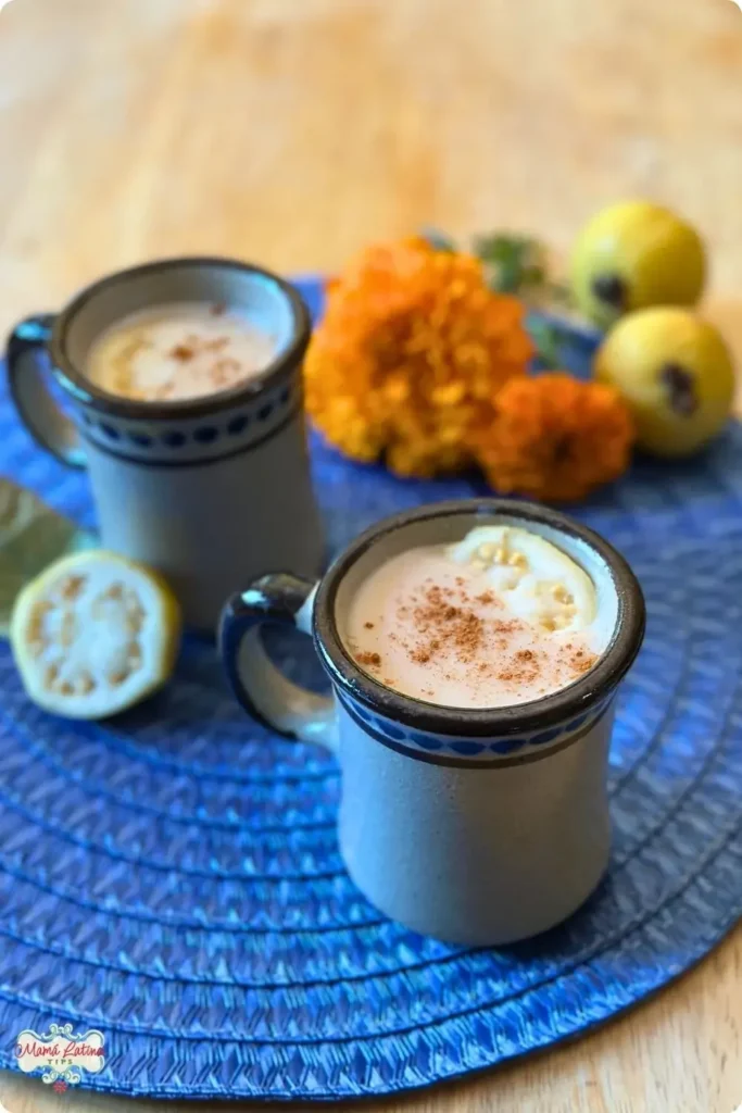 Dos tazas de cerámica mexicana con atole de guayaba adornadas con canela en polvo y una rodaja de guayaba encima. Las tazas están sobre un mantel redondo de paja en color azul y sobre una mesa de madera. Alrededor de las tazas hay guayabas enteras y cortadas por mitad y varias flores de cempasúchil 
