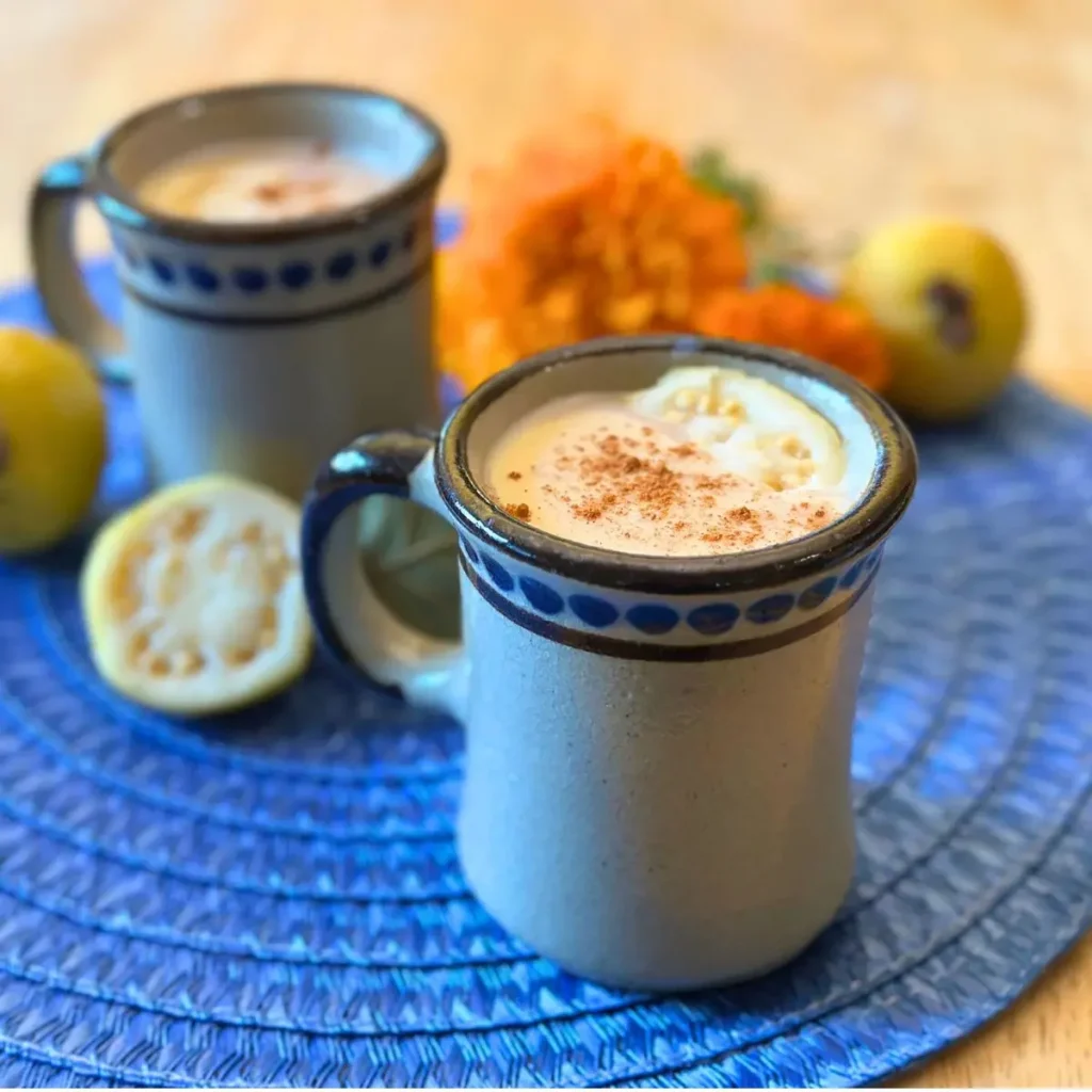 Two ceramic mugs filled with a creamy Guava Atole, topped with cinnamon, sit on a blue woven mat with cut guavas and orange flowers in the background.