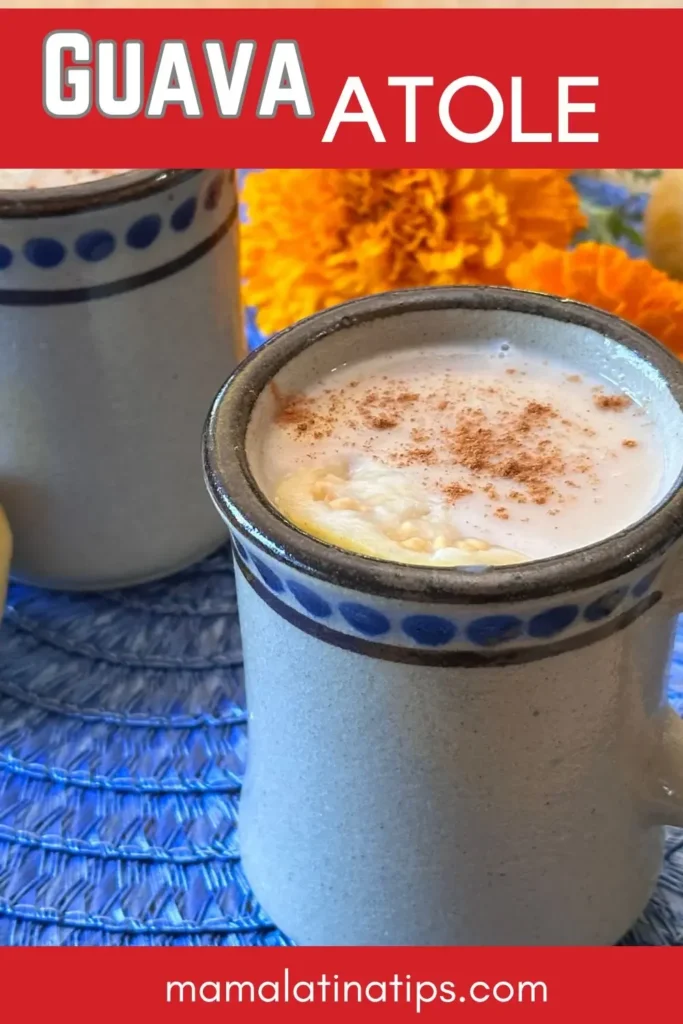 Two ceramic mugs filled with homemade Guava Atole Recipe, topped with cinnamon, sit on a blue woven mat with marigold flowers in the background.