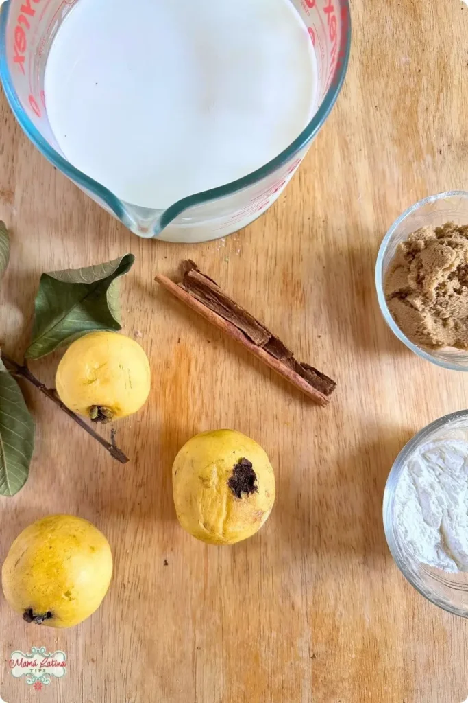 Una tabla medidora con leche, tres guayabas amarillas con hojas verdes a un lado. Una raja de canela y dos tazones chiquitos, uno con azúcar morena y otro con fécula de maíz. Ingredientes para preparar atole de guayaba, sobre una mesa de madera. 