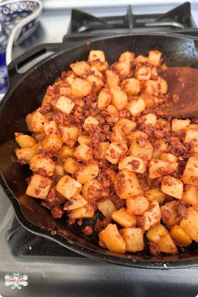 A cast iron skillet on a stove contains diced potatoes and Mexican Chorizo being cooked together for a classic papas con chorizo dish.