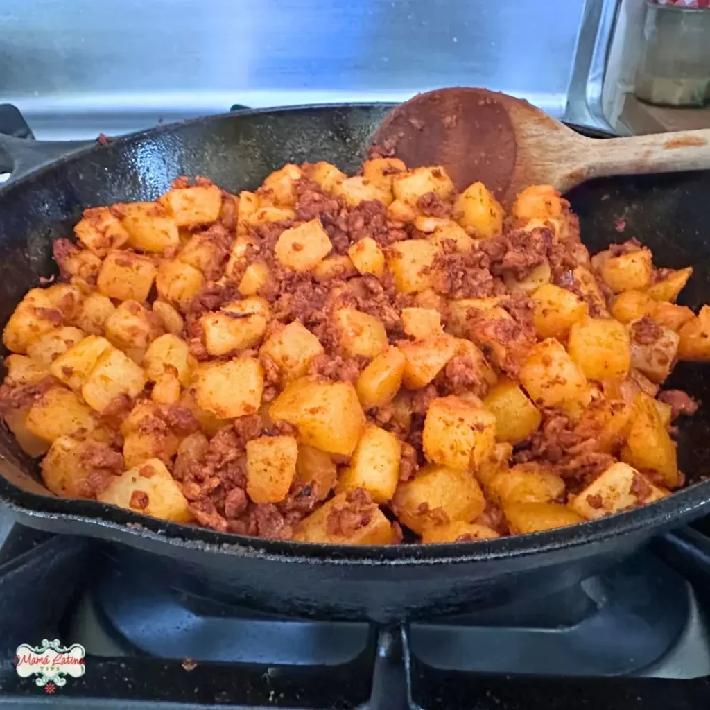 A cast iron skillet on a stovetop filled with cooked diced potatoes and Mexican Chorizo, stirred with a wooden spoon.