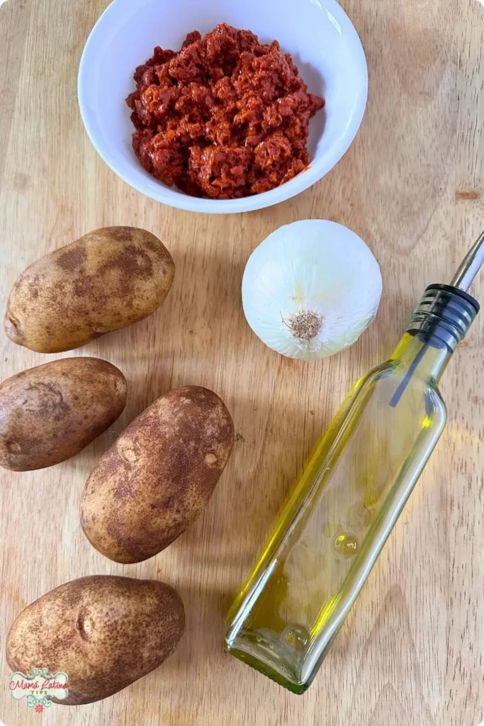 Four potatoes, a whole white onion, a bottle of olive oil, and a bowl of chopped Mexican Chorizo are arranged on a wooden surface