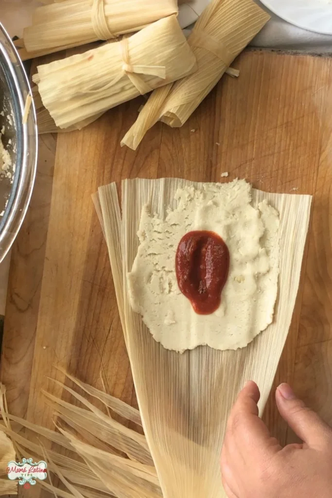 Una mano extiende masa y salsa roja sobre una hoja de maíz para preparar tamales. Cerca, tamales atados descansan sobre una superficie de madera, una escena atractiva para cualquiera que siga una guía paso a paso para preparar hojas de tamal.