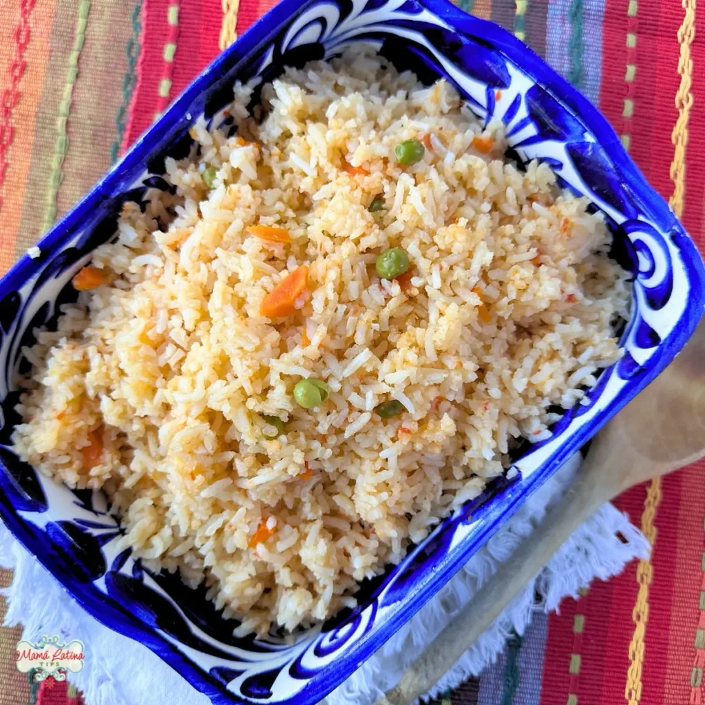 A rectangular blue and white bowl filled with Mexican rice containing peas and carrots, placed on a colorful striped cloth with a wooden spoon nearby.
