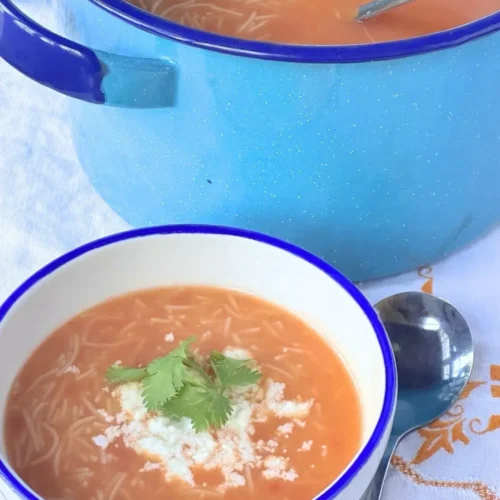 A bowl of Mexican fideo soup topped with cheese and cilantro sits next to a spoon, with a large blue pot of more soup in the background.