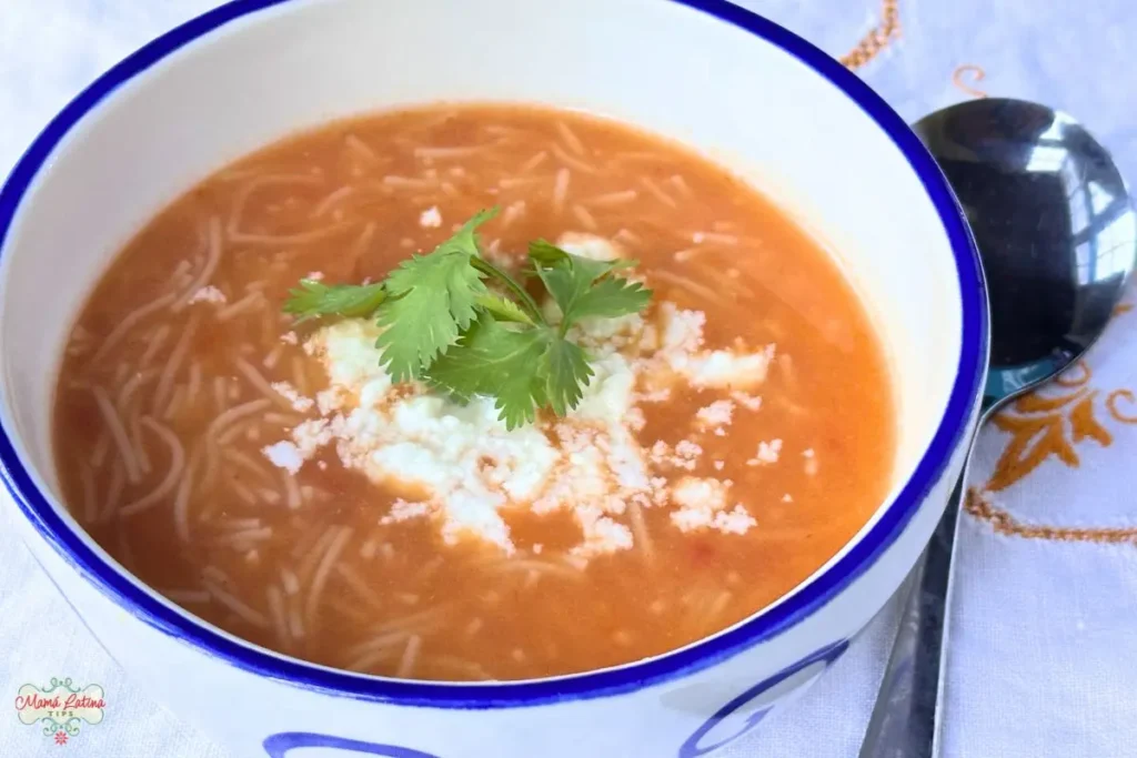 A bowl of Mexican fideo soup, garnished with crumbled cheese and fresh cilantro, sits on a white tablecloth with a spoon beside it.