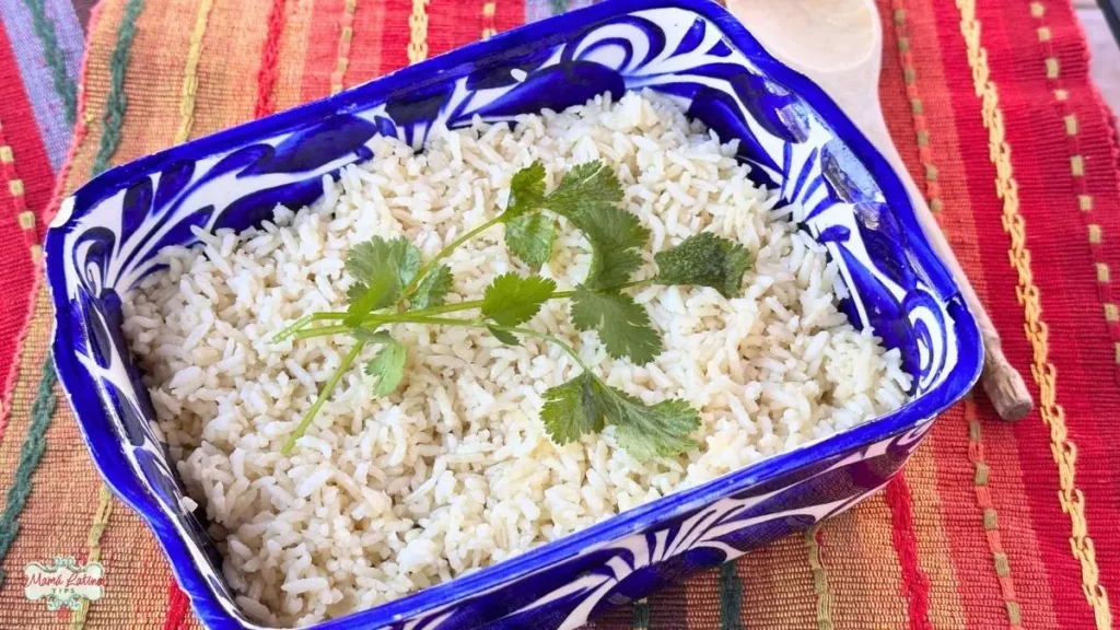 A blue patterned dish filled with cooked Mexican White Rice, garnished with fresh cilantro, sits on a colorful woven tablecloth with a wooden spoon beside it.