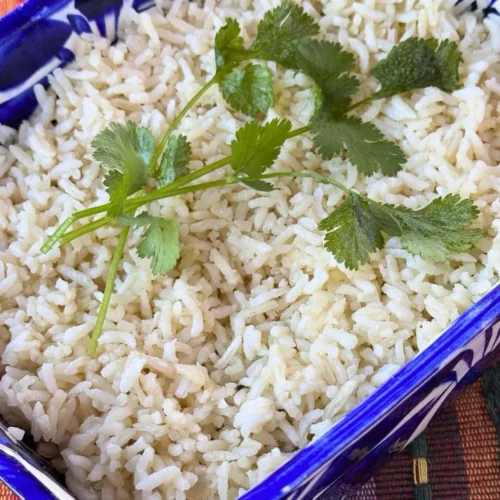 A rectangular blue and white dish filled with cooked Mexican White Rice, garnished with sprigs of cilantro, sits on a colorful woven tablecloth.