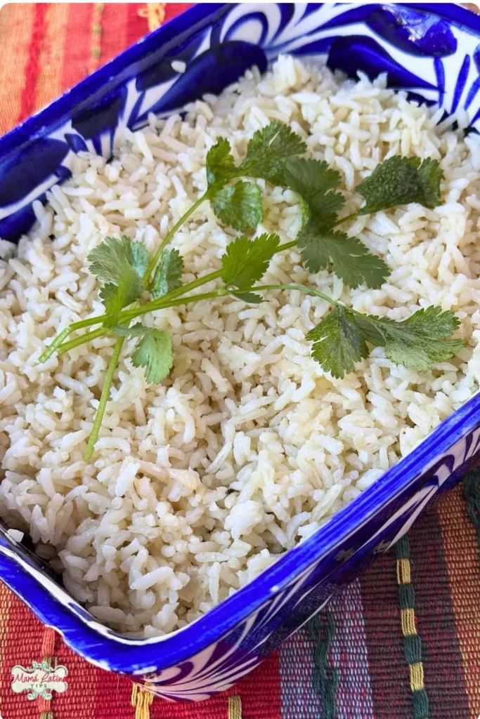 A rectangular blue and white dish filled with Mexican White Rice, garnished with sprigs of fresh cilantro, sits on a colorful woven tablecloth.