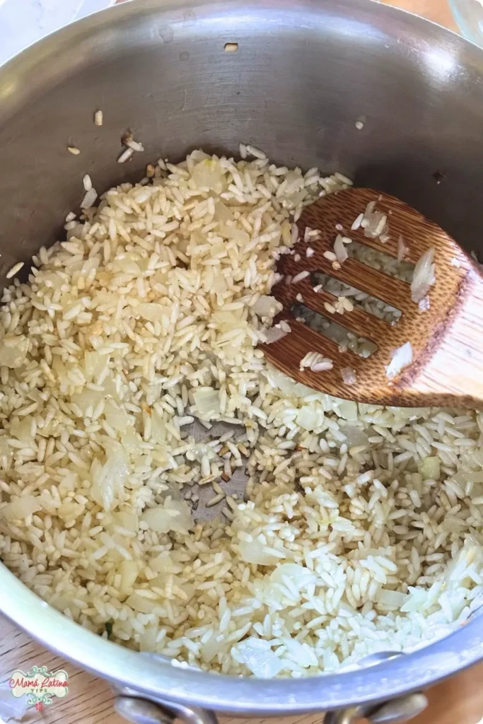 Uncooked White Rice and chopped onions being stirred with a wooden spoon in a stainless steel pot, the first steps to preparing delicious Mexican White Rice.