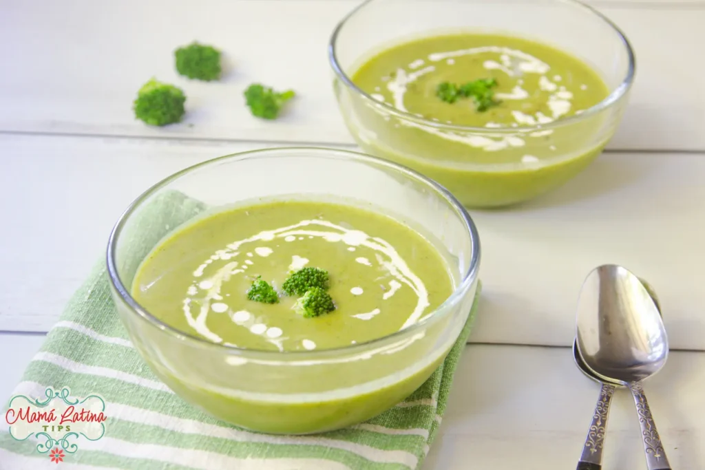 Two bowls of Cream of Broccoli Soup, garnished with broccoli florets and a drizzle of cream, sit on a striped cloth next to two spoons.