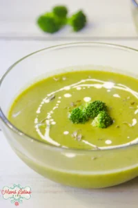 A clear glass bowl of Cream of Broccoli Soup with a swirl of cream and broccoli florets on top, set on a white surface with broccoli pieces in the background.