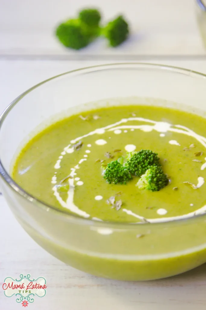 A clear glass bowl of Cream of Broccoli Soup with a swirl of cream and broccoli florets on top, set on a white surface with broccoli pieces in the background.
