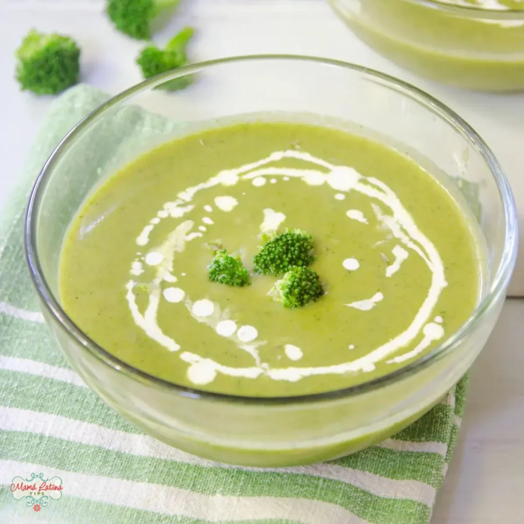 A glass bowl of Cream of Broccoli Soup, garnished with a swirl of cream and small broccoli florets, sits atop a green and white striped cloth.