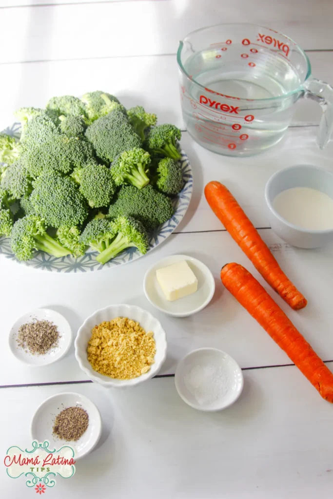 Ingredients for Cream of Broccoli Soup displayed on a white surface: broccoli florets, two carrots, a measuring cup of water, butter, seasonings, and a small bowl of cheese powder.